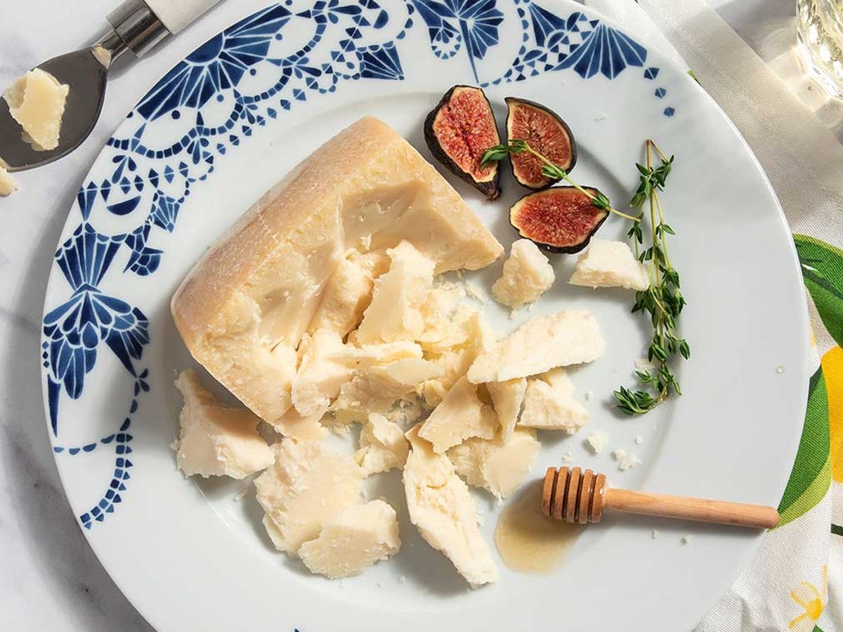A rinded wedge of aged sheep cheese broken into craggy chunks on a blue and white plate with halved figs, thyme and a honey dipper