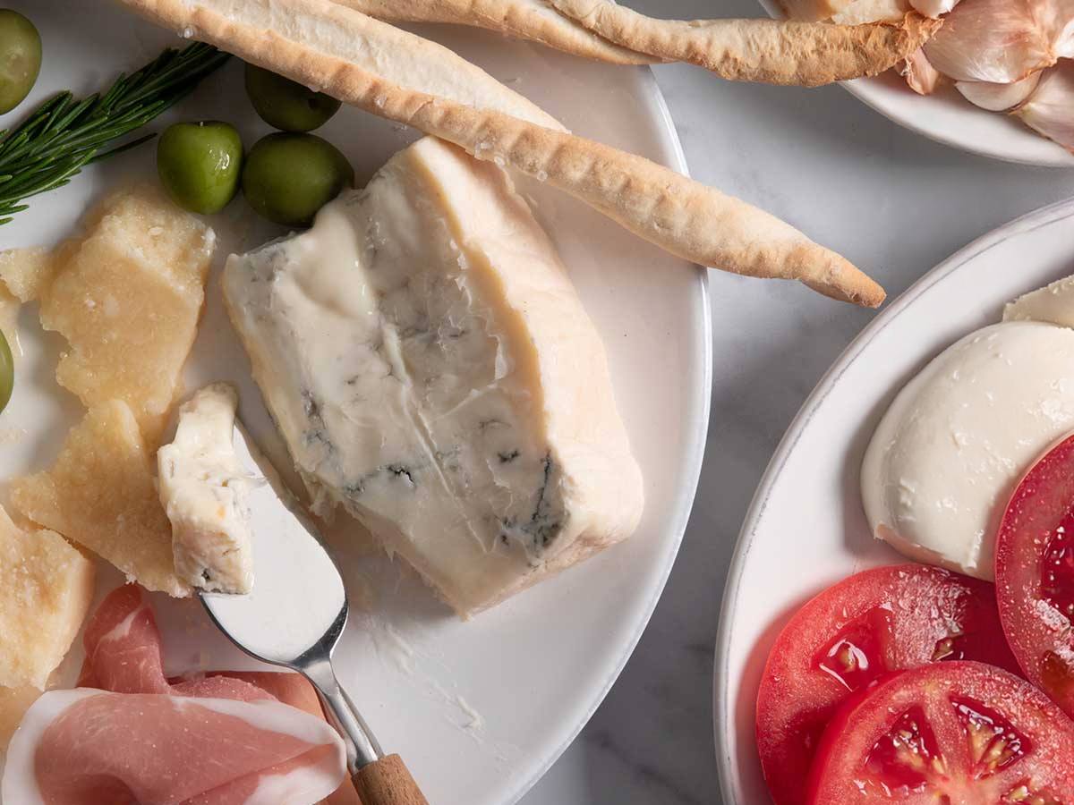 A creamy blue-veined wedge of Gorgonzola Dolce on a plate with green olives, grissini, prosciutto and shards of hard grating cheese