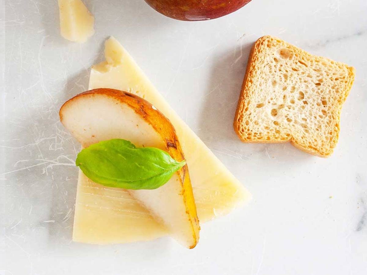 A thin wedge of pale aged pecorino topped with a slice of roasted pear and a basil leaf, beside a slice of toast