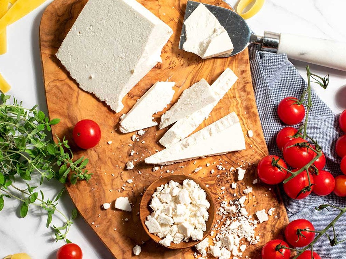A block of ricotta salata on an olive wood board, cut into wedges and crumbled into a small bowl, with vine cherry tomatoes, fresh oregano and dried pasta